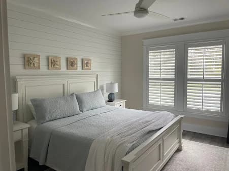 Interior painting project by Of The Essence Design Build Paint in Wilmington, NC. The image shows a light bedroom with white trim and shuttered windows.