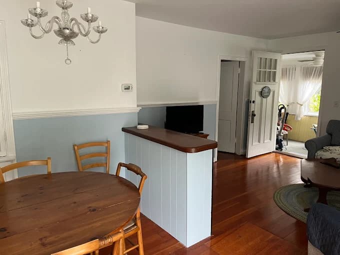 This is an image from CertaPro Painters of Cape Cod, located in Massachusetts. The photo shows a cozy dining and living space with freshly painted two-tone walls in white and light blue, complemented by wood flooring and natural light.