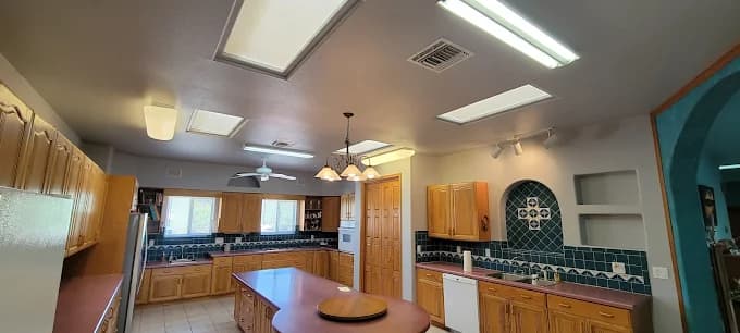 This is an image from Innovative Coatings LLC, located in New Mexico. The photo shows a spacious kitchen with natural wood cabinets, teal tile backsplash, recessed lighting, and a central island beneath skylights and ceiling fixtures.