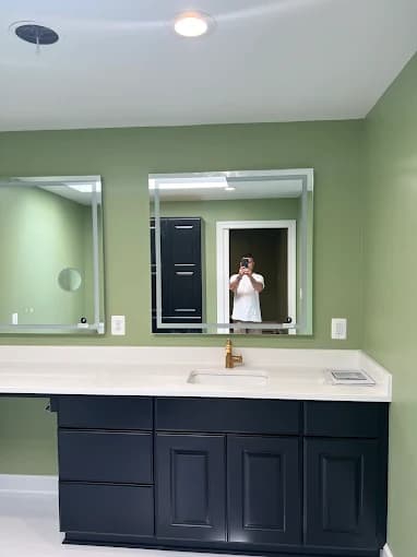 This is an image from Mendez Drywall & Painting LLC, located in Maryland. The image shows a bathroom vanity with dark blue cabinets, a white countertop, gold faucet fixtures, and soft green painted walls.