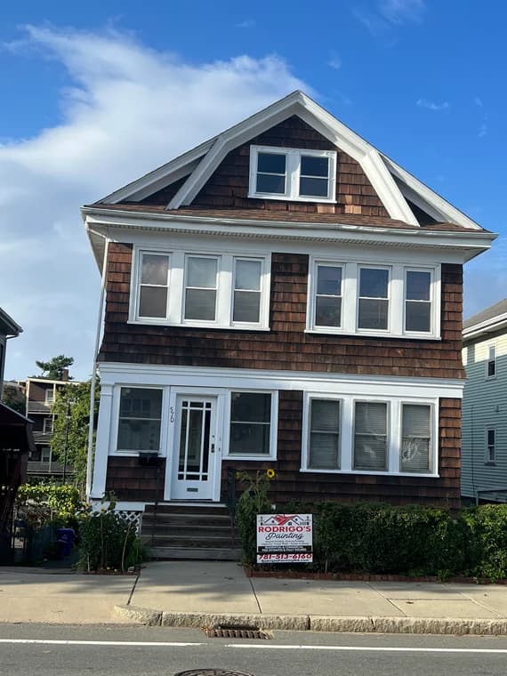 This is an exterior house painted by Rodrigo's Painting. The multi-story home has natural brown shingle siding with white trim, and the company’s lawn sign is displayed in front.