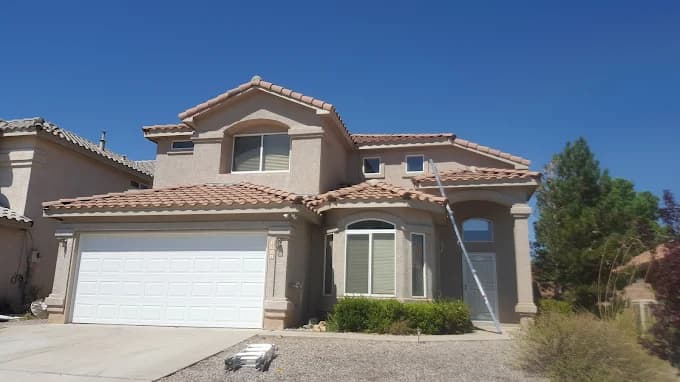 This is an image from Mike’s Pro Painting, LLC, located in New Mexico. The photo shows a two-story stucco home with a tile roof being painted, with a ladder positioned against the wall and bright desert landscaping under a clear blue sky.