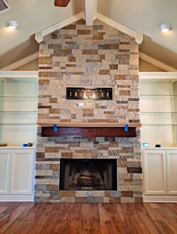 "This is an image from Prestige Pro Painters, located in Texas. The photo shows a stone-faced living room fireplace with built-in shelving, a wooden mantel, and a mounted TV bracket above the firebox."