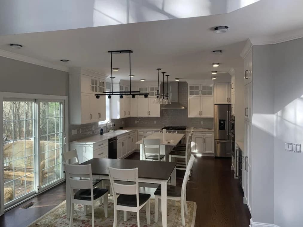 This is an image from Hoffman Interior Painting, located in Michigan. The image shows a freshly painted modern kitchen interior with white cabinetry, stainless steel appliances, gray backsplash tile, recessed lighting, and clean light gray walls demonstrating professional interior and cabinet painting services.