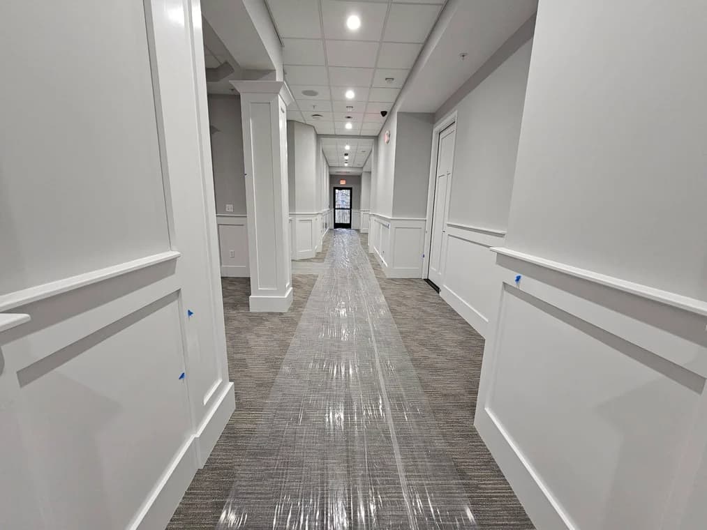 This is an image from Great Lakes 1-800 Painting LLC company, located in Davison, MI The image is a freshly painted commercial hallway with white wainscoting, soft gray walls, and protective plastic covering over carpet — interior painting by Great Lakes Painting.