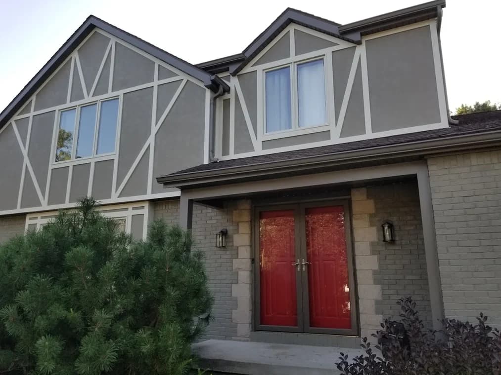 This is an image from Queen City Painting & Wash, located in Ohio. The photo shows the same Tudor-style home after exterior painting, featuring updated gray stucco, crisp white trim, and red double entry doors.