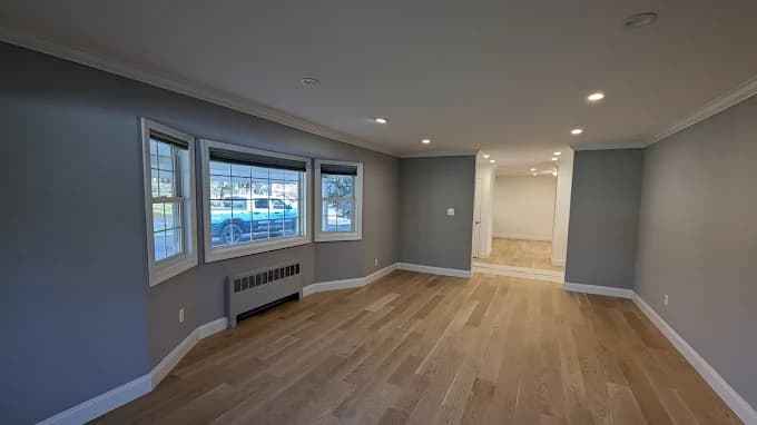 This is an image from Precision Painting Plus of Suffolk County, located in New York. The photo shows a freshly painted open room with gray walls, recessed lighting, and new light wood flooring.