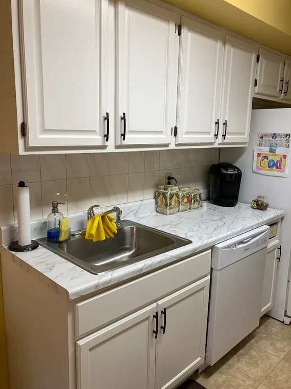 This is an image from CertaPro Painters of Staten Island, located in New York. The photo shows a kitchen with freshly painted white cabinets, black hardware, a stainless steel sink, and marble-style countertops.