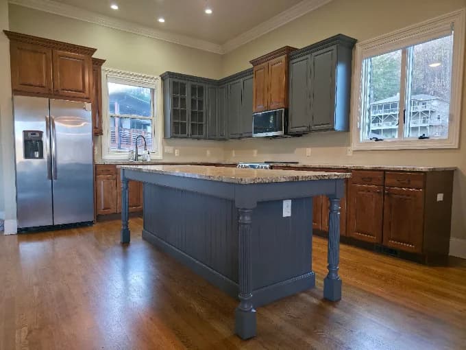 Interior cabinet painting project by Buchanan Painting in Hendersonville, NC. The image shows a kitchen with gray island panels, wood cabinets, and windows.