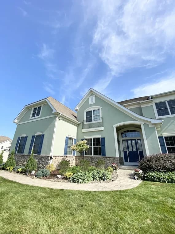 This is an image from KB Quality Painting, located in New York. The image shows a large two-story home with light green exterior walls, blue shutters, and detailed stonework along the lower facade.