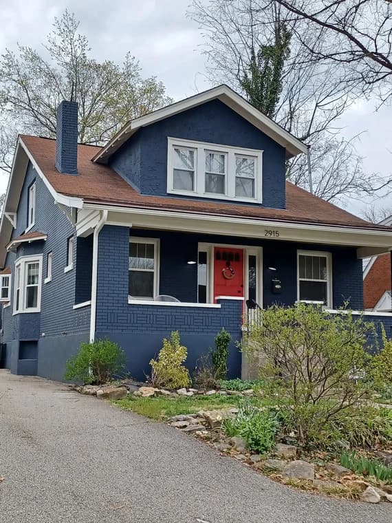 This is an image from Abell Painting LLC, located in Kentucky. The image shows a two-story brick home painted in navy blue with white trim and a bright red front door, showcasing a refreshed exterior finish.