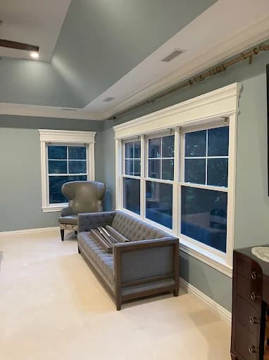 This is an image from Jay's Pro Painting, located in New York. The photo shows a softly lit sitting room with blue-gray walls, white trim, and a tufted gray loveseat positioned beneath wide double windows.