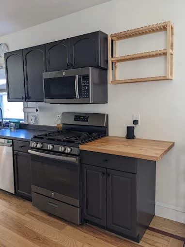 This is an image from Pro Painting Plus, located in New York. The photo shows a modern kitchen with dark painted cabinets, stainless steel appliances, a butcher-block countertop extension, and a light wood floor.