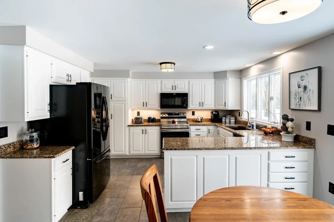 This is an image from A&L Painting, located in New York. The photo shows a bright kitchen featuring repainted white cabinets, granite countertops, and black appliances.