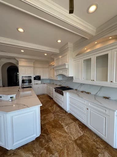 This is an image from Precision Painting Plus of Suffolk County, located in New York. The photo shows a newly refinished white kitchen with detailed cabinetry, marble countertops, and warm recessed lighting.