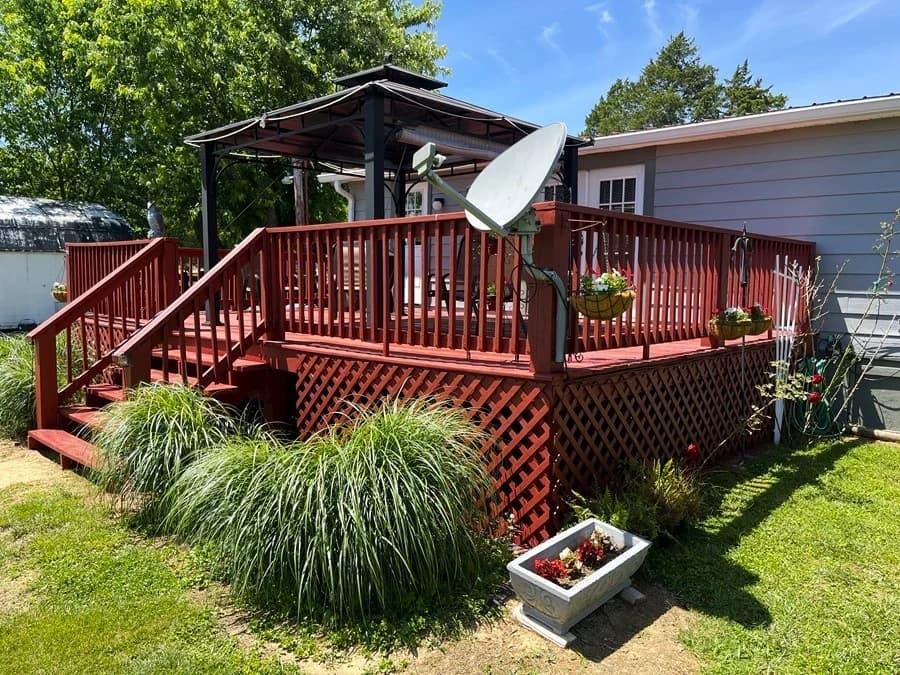 This is an image from Quick Painting LLC, located in Kentucky. The image shows a red-painted outdoor deck with railings, lattice skirting, and stairs, surrounded by landscaped greenery and outdoor seating.