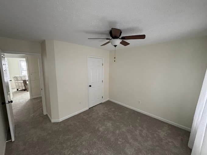 This is an image from Bates Brothers Painting LLC, located in New York. A cleanly painted bedroom with smooth beige walls, white trim, brown carpeting, and a ceiling fan, showcasing a professional interior paint finish.