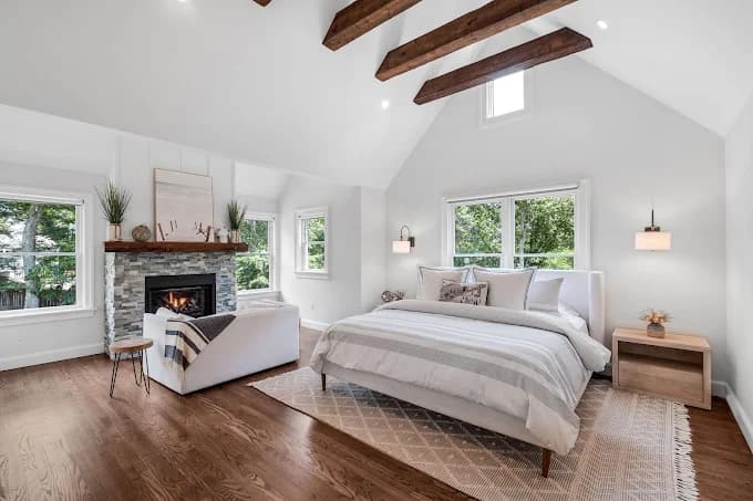This is an image from Chameleon Painting Inc., located in New York. The photo shows a bright, modern bedroom with white walls, exposed ceiling beams, a stone fireplace, hardwood floors, and large windows bringing in natural light.