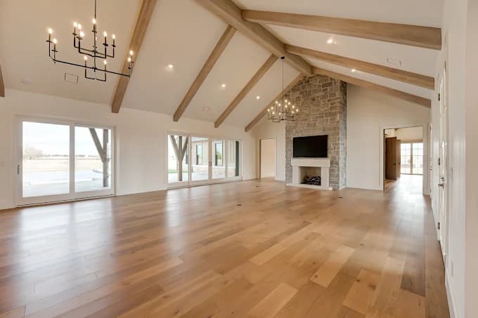 This is an image from CertaPro Painters of OKC Edmond, located in Oklahoma. The photo shows a spacious living room with vaulted ceilings, exposed wooden beams, a stone fireplace with mounted TV, and wide-plank hardwood floors.