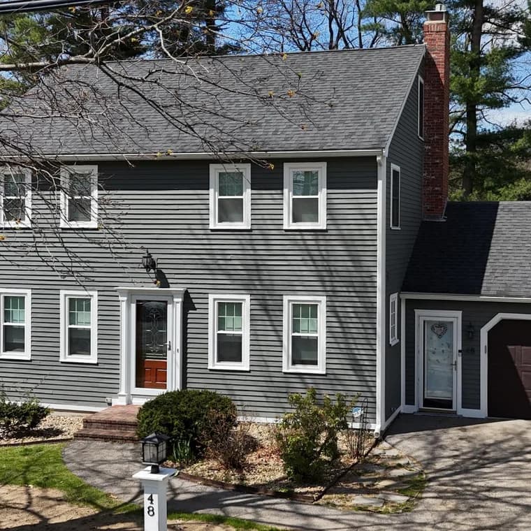 This is an image from Paint Bros MA, located in Massachusetts. It shows a freshly painted gray two-story home with white trim and a brick chimney, showcasing the company’s professional exterior painting services.