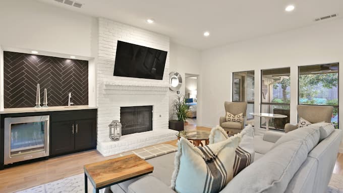 This is an image from Big Al's Painting, located in Texas. The photo shows a bright living room with a white painted brick fireplace, a mounted TV, black wet bar cabinetry with herringbone tile, and large windows overlooking the backyard.
