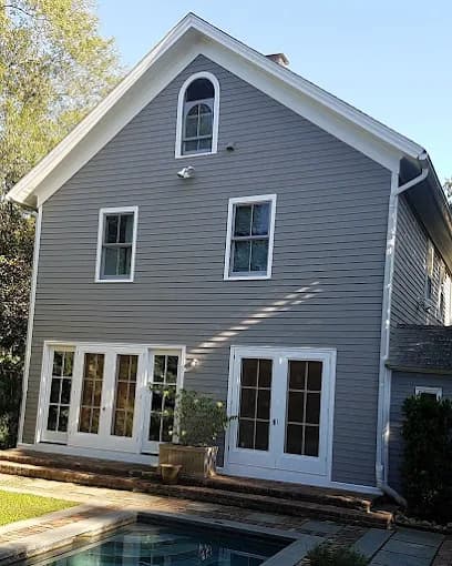This is an image from Chameleon Painting Inc., located in New York. The photo shows a gray two-story home exterior with white trim and multiple windows, freshly painted and overlooking a poolside patio.