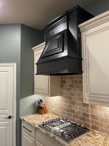 This is an image from MJ Workforce Solutions LLC, located in Texas. The kitchen view highlights a black custom wood vent hood above a gas cooktop with cream cabinetry and a brick-style backsplash.