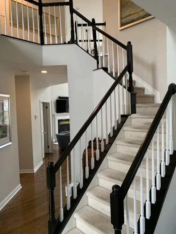 This is an image from Hoffman Interior Painting, located in Michigan. The image shows a freshly painted interior staircase with white balusters, black handrails, beige carpeted steps, and clean light gray walls highlighting professional interior painting and trim finishing services.