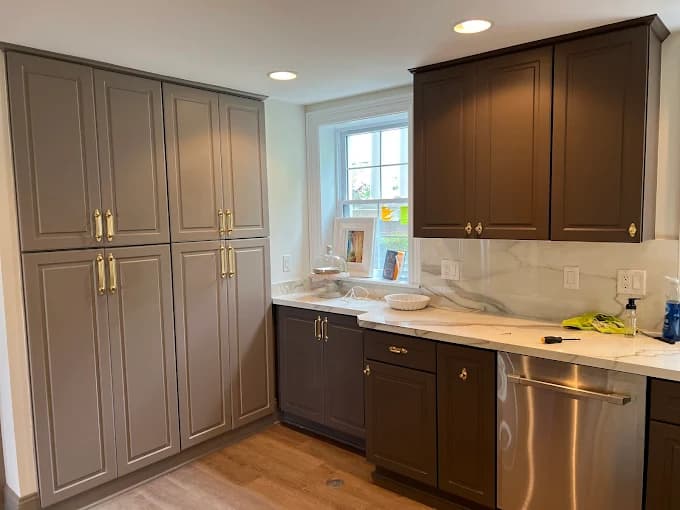 This is an image from Advantage Paint Services, located in New York. The image shows freshly painted two-tone kitchen cabinets with gray and dark brown finishes, gold hardware, and a marble-look countertop.