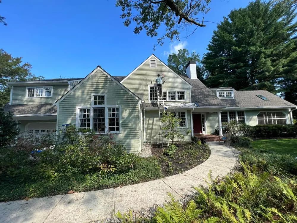Large light green house with multiple windows and a gray roof, where a painter is on a ladder applying fresh paint to the siding, representing exterior painting services by Painting Plus Home Improvement LLC.