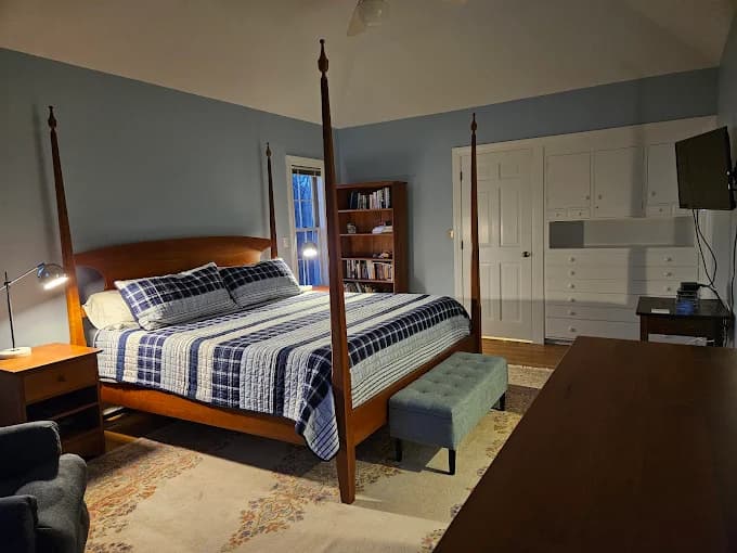 This is an image from Stewart Painting, located in Massachusetts. The photo shows a bedroom with soft blue painted walls, white built-in cabinetry, and a wooden four-poster bed with plaid bedding.