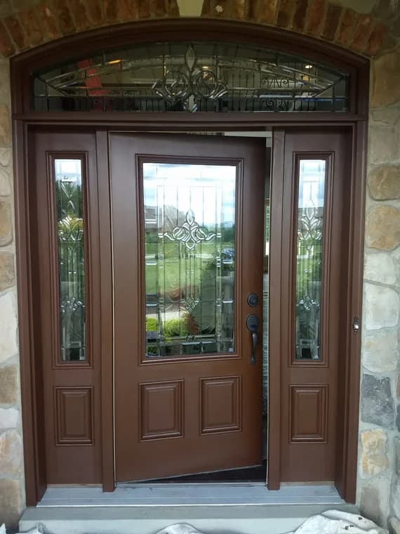 This is an image from Roman Paint Pros company, located in Penn Hills, PA. The image is a freshly painted brown front entry door with decorative glass panels and sidelights, framed by stone and brick exterior, professionally refinished by Roman Paint Pros.
