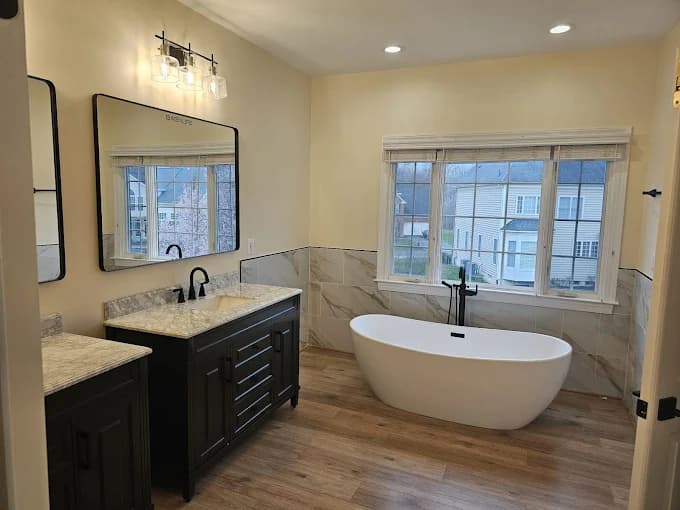 This is an image from Julio's Painting LLC, located in Maryland. The photo shows a remodeled bathroom with a freestanding tub, dual vanities, large mirrors, and light wood flooring.