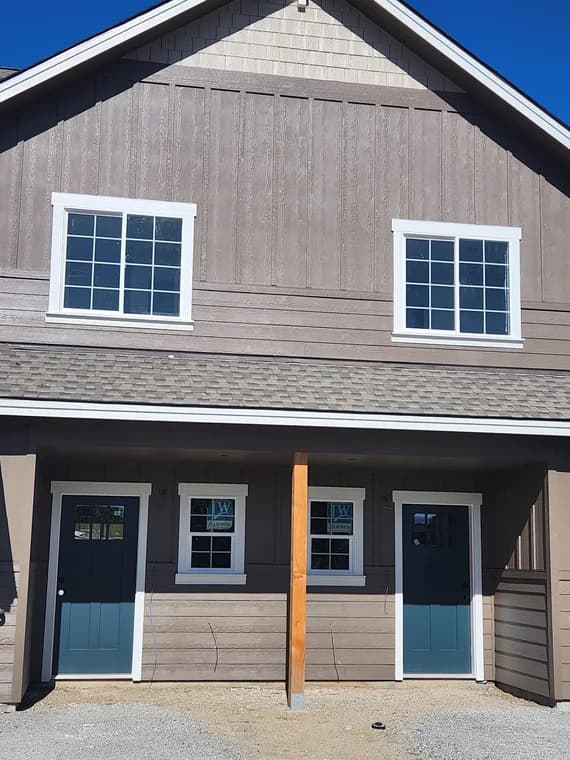 This is an image from Honey Do Painting LLC company, located in Bonners Ferry, ID The image is a freshly painted two-story duplex with taupe siding, white window trim, blue front doors, and shake siding in the gable, exterior painting by Honey Do Painting