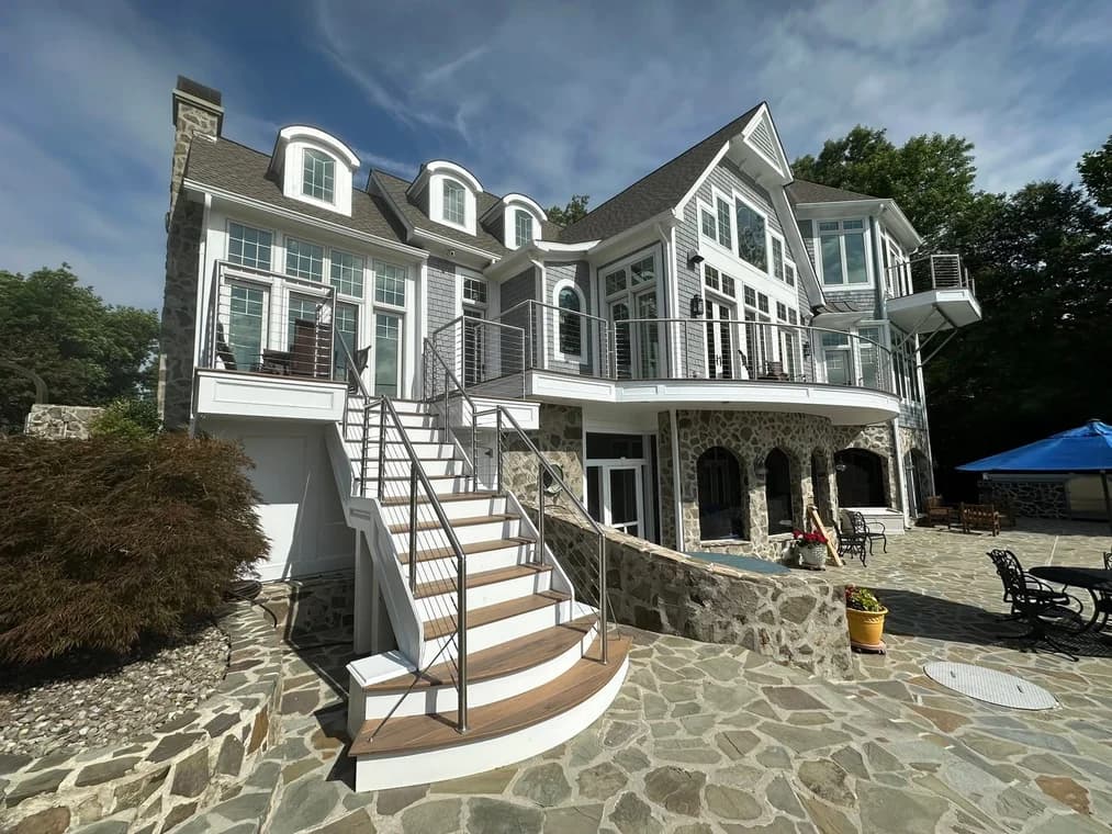This is an image from DJ’s Painting & Powerwashing, located in Maryland. The photo shows a large multi-story stone and siding home with white trim, expansive windows, and a decorative curved patio area.