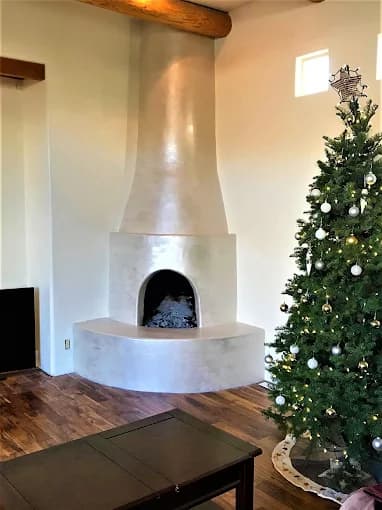 This is an image from Busby Finishes, located in New Mexico. The photo shows an elegant living room with a smooth plaster kiva-style fireplace, wooden ceiling beams, and a decorated Christmas tree beside it.