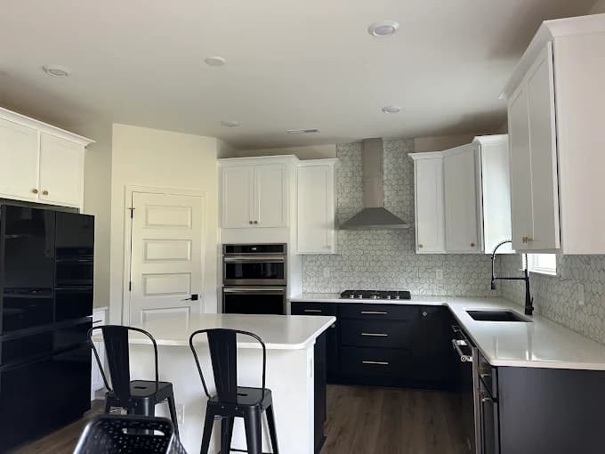 This is an image from 2 Cabinet Girls, located in North Carolina. The photo shows a remodeled kitchen with white upper cabinets, black lower cabinets, a white quartz countertop, and a stainless steel range hood creating a sleek modern contrast.