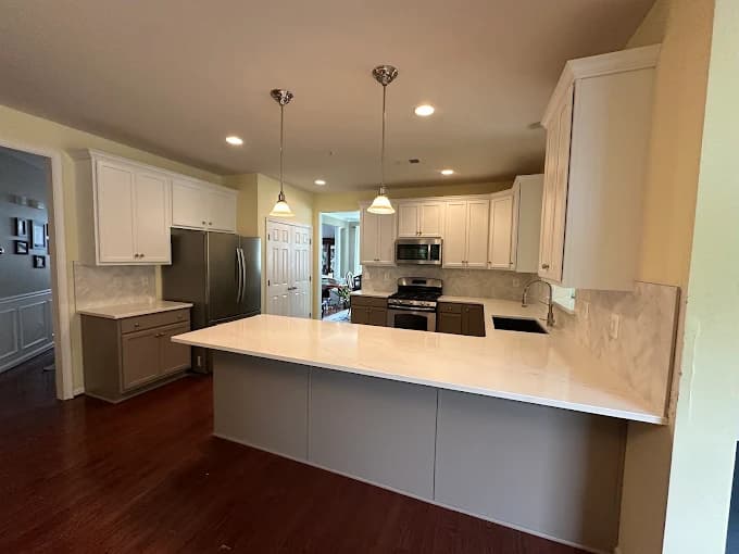 This is an image from C&R Pro Painting, located in Virginia. The photo shows a modern kitchen with freshly painted white upper cabinets, gray lower cabinets, stainless steel appliances, and a bright quartz countertop.