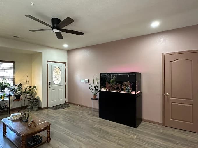 This is an image from New Mexico's Painting Specialists, located in New Mexico. The photo shows a freshly painted entryway and living space with mauve and cream-colored walls, a black aquarium, a ceiling fan, and wood-look tile flooring.