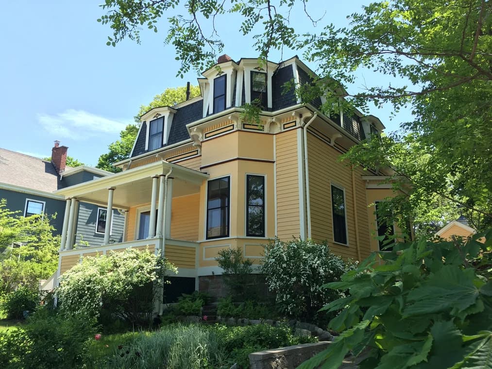 This is an image from Catchlight Painting, located in Massachusetts. It shows a yellow Victorian-style home with black roof shingles and white trim, surrounded by greenery and trees.