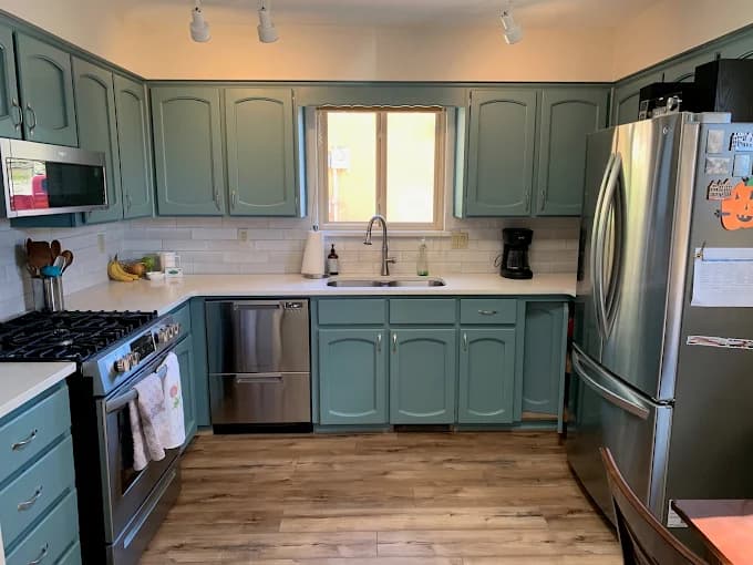 This is an image from Straight Line Painting LLC, located in New Mexico. The photo shows a remodeled kitchen with teal-painted cabinets, white countertops, stainless steel appliances, and wood-look flooring.