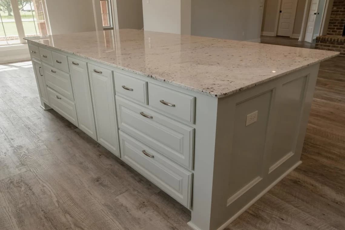 This is an image from Schmidt Painting, located in Massachusetts. It highlights a kitchen island refinished by Schmidt Painting, with light-colored cabinetry, silver handles, and a granite countertop set on light wood flooring.