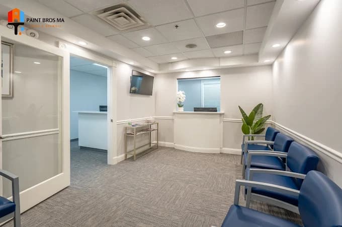 This is an image from Paint Bros MA, located in Massachusetts. The photo shows a freshly painted office waiting area with soft gray walls, white trim, recessed lighting, and blue seating, creating a clean and professional atmosphere.