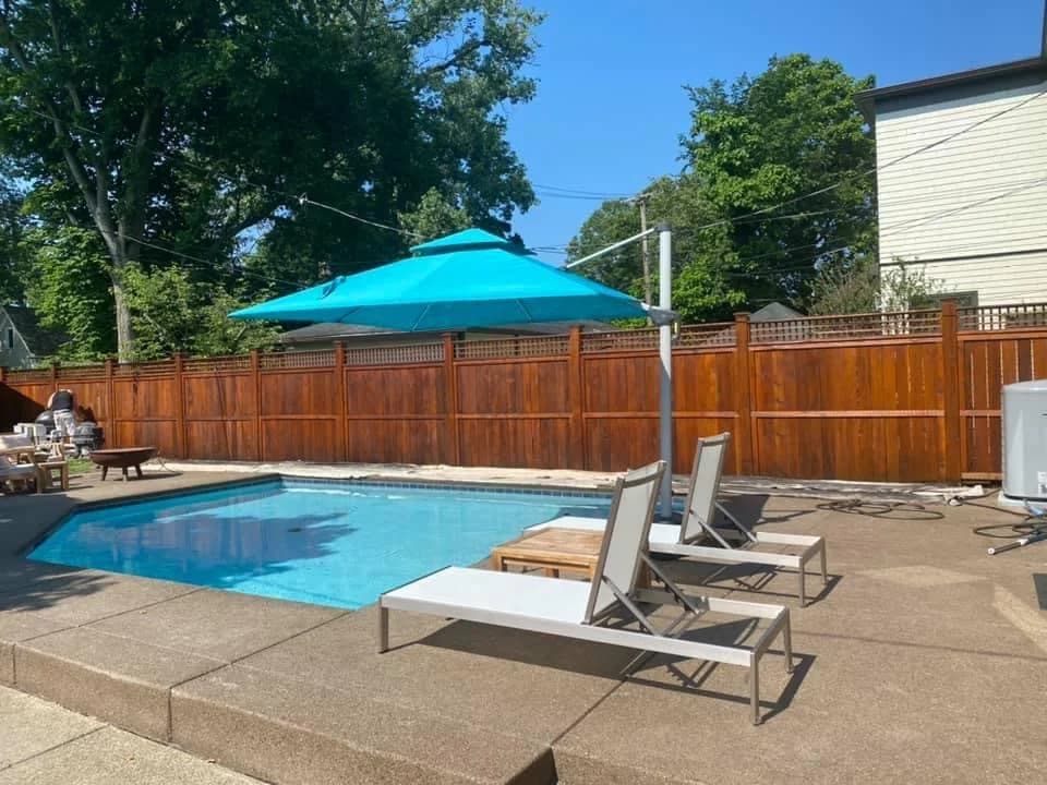 This is an image from Queen City Painting & Wash, located in Ohio. The outdoor pool area is shown with a freshly stained wooden privacy fence surrounding a clean concrete patio and lounge chairs.