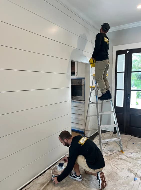 This is an image from Bright Side Painting Of Jax company, located in Jacksonville Beach, FL. The image is an interior house painters from Bright Side Painting applying fresh white paint to shiplap wall in modern home using brush and ladder