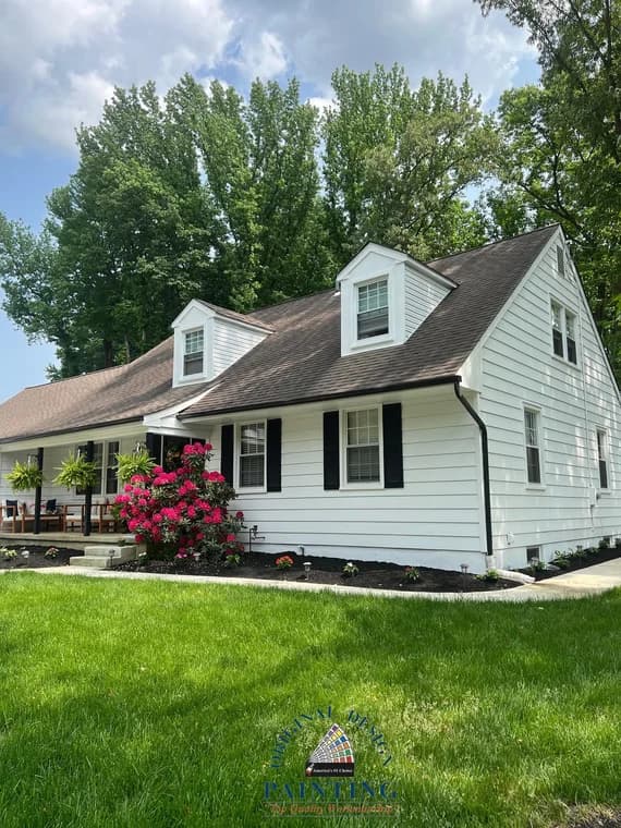 This is an image from Original Design Painting, highlighting a white Cape Cod–style home with black shutters. The lawn is neatly landscaped with flowering shrubs and trees.