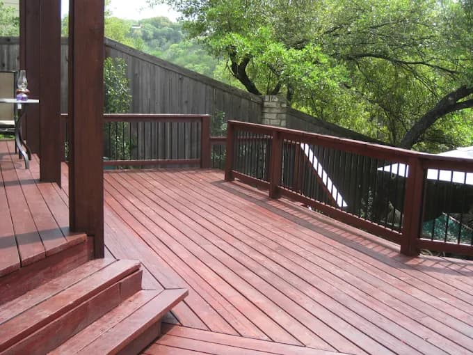 This is an image from Next Door Painting, located in Texas. The photo shows a freshly stained wooden deck with rich red-toned boards and matching railings surrounded by a backyard with trees and fencing.
