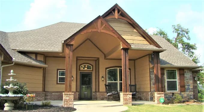 This is an image from Hometown Painting, located in Oklahoma. The photo shows the exterior of a tan and brown Craftsman-style home with wood accents, brick details, and a welcoming front porch with rocking chairs.