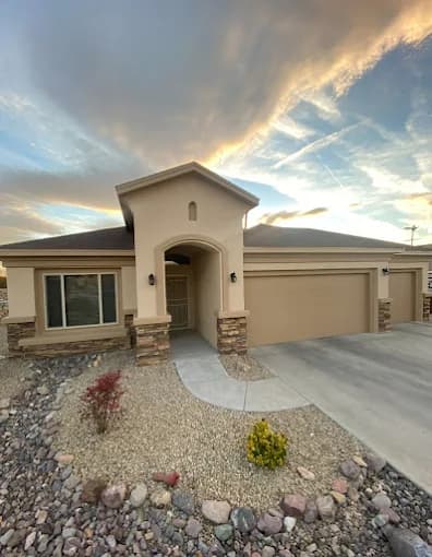 This is an image from Innovative Coatings LLC, located in New Mexico. The photo shows a modern tan stucco home with stone accents, a neatly landscaped gravel front yard, and a wide driveway under a colorful evening sky.