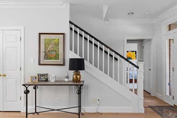 This is an image from Gonzalez Painters & Contractors, Inc., located in North Carolina. The photo shows a bright interior entryway with freshly painted white walls, a staircase with black handrails, light wood flooring, and a decorative console table.